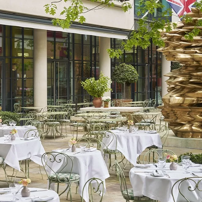 Outdoor restaurant with wrought iron tables and chairs, each set with white tablecloths and place settings. There are potted plants, large windows, and a golden abstract sculpture on the right. Greenery and a British flag are visible above.