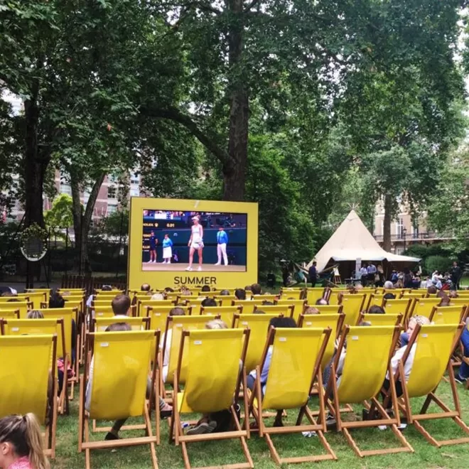 Rows of yellow deck chairs are set up on grass facing a large outdoor screen showing a tennis match. People are seated, and tall trees and a tent are in the background, creating a summery park setting.