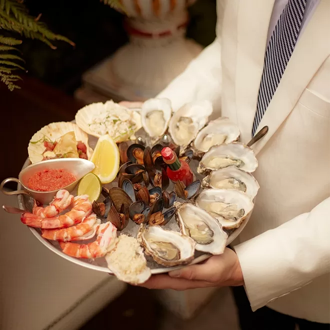 A person in a white suit holds a platter of assorted seafood, including oysters, mussels, shrimp, lemon wedges, cocktail sauce, and a small bottle of hot sauce. The platter is arranged neatly and looks fresh.