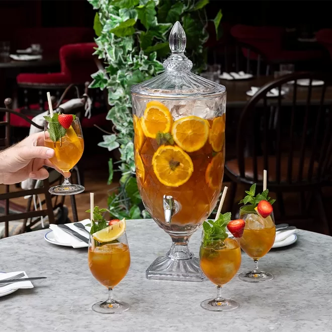 A glass beverage dispenser filled with iced tea, orange slices, and mint sits on a marble table, surrounded by four garnished glasses, with one hand reaching for a drink. Greenery and dining chairs are visible in the background.