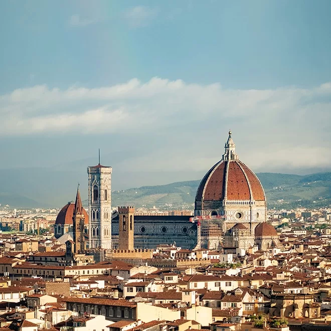 A panoramic view of Florence, Italy, featuring the iconic Florence Cathedral with its large red-tiled dome, bell tower, and surrounding city buildings under a clear sky.