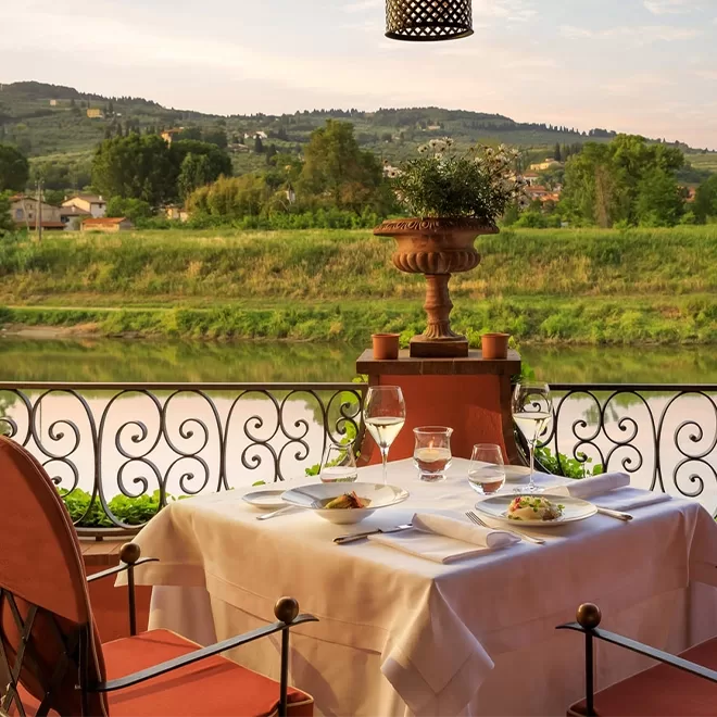 A table set for two with white linens and elegant place settings overlooks a river and lush green hills, surrounded by an ornate balcony railing on an outdoor terrace.