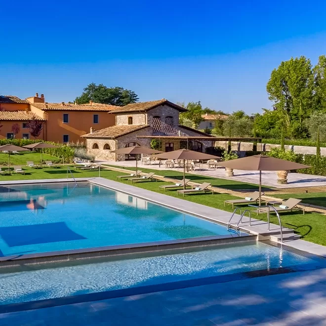 A large outdoor swimming pool surrounded by lounge chairs and umbrellas sits in front of rustic stone and stucco buildings under a clear blue sky, with green lawns and trees in the background.