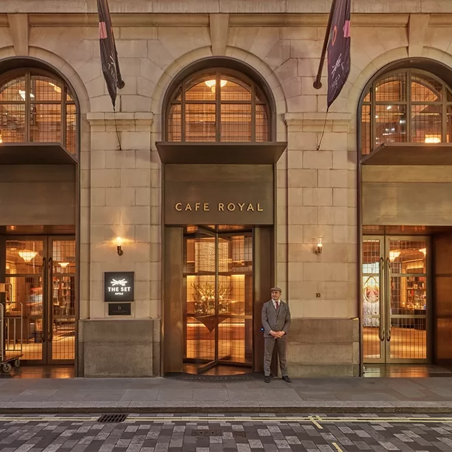 The entrance of Café Royal, featuring large arched windows, warm interior lighting, and a doorman in a suit standing by the revolving door on a cobblestone street.