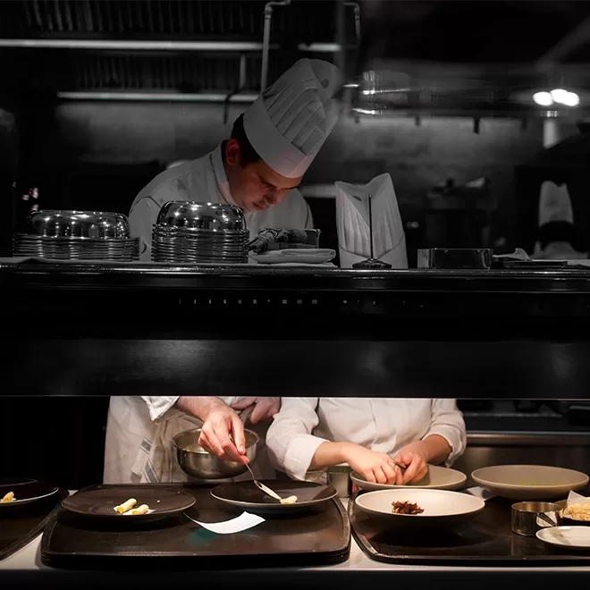 A chef in a white uniform and hat carefully plates food in a dimly lit restaurant kitchen, while another person assists beside him, partially out of view behind a counter.