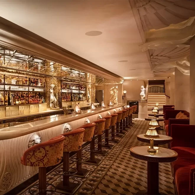 Luxurious bar interior with ornate decor, a long marble counter lined with plush, upholstered stools, small tables with lamps, golden accents, and warm, ambient lighting creating an elegant atmosphere.