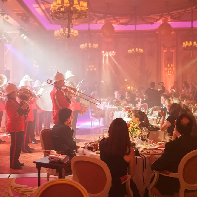 A brass band in red uniforms performs in a lavish, warmly lit ballroom as elegantly dressed guests sit at round tables, dining and watching the entertainment. Chandeliers and ornate decor add to the festive atmosphere.