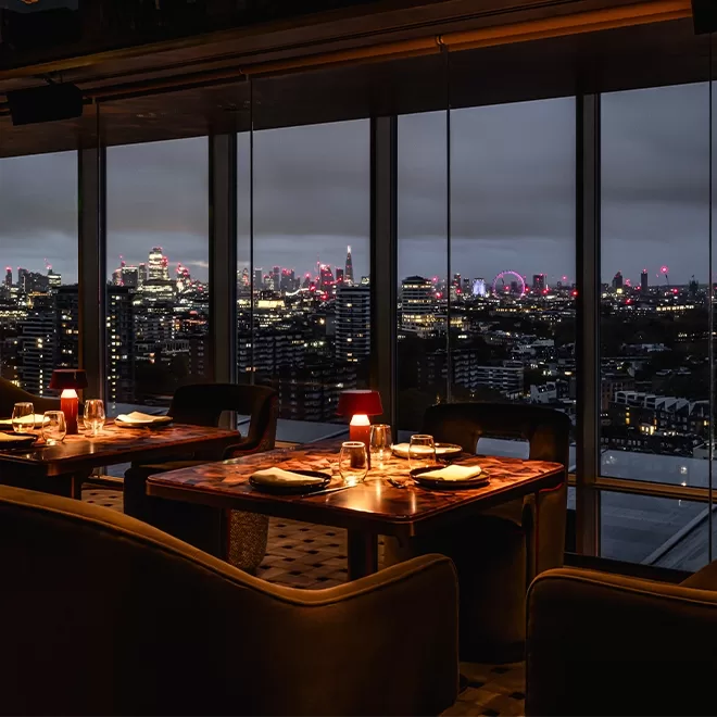Elegant restaurant with set tables and warm lighting, featuring floor-to-ceiling windows that reveal a city skyline at dusk, including illuminated skyscrapers and the London Eye in the distance.