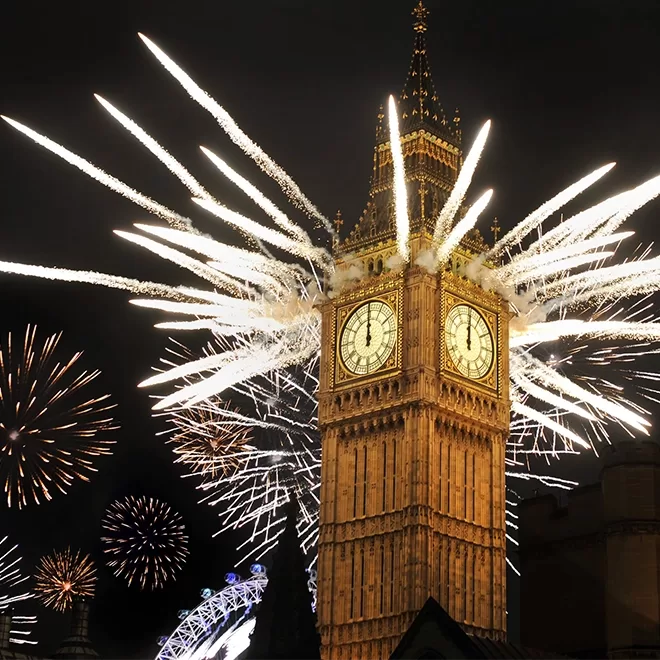 Big Ben clock tower at night with bright fireworks exploding around it and the London Eye visible in the background. The sky is filled with colorful sparks and trails of light.