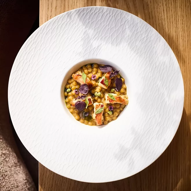 A white textured plate holds a gourmet dish of creamy, golden risotto garnished with small pieces of fish, purple microgreens, and herbs, presented on a light wooden table.