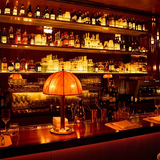A dimly lit bar with shelves stocked with bottles of liquor, glassware, and a glowing amber lamp on a polished wooden counter. Glasses, cocktail tools, and napkins are neatly arranged on the bar.