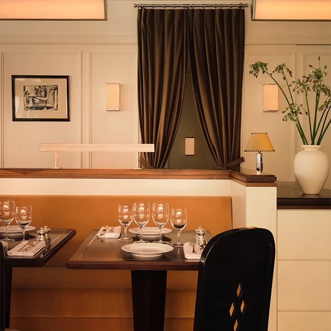 A cozy restaurant interior with tan leather seating, brown curtains, set tables with glassware, a framed picture on the wall, and a large white vase with flowers on a counter. Warm lighting creates an inviting atmosphere.