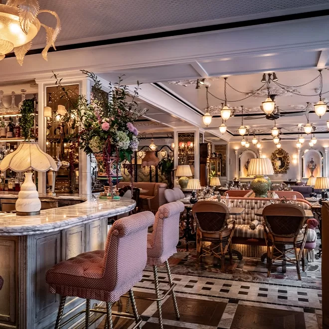 A cozy, elegant restaurant interior with plush pink bar stools, a marble countertop, vintage lamps, floral arrangements, and warm lighting. The space features cushioned seating, ornate decor, and a welcoming ambiance.