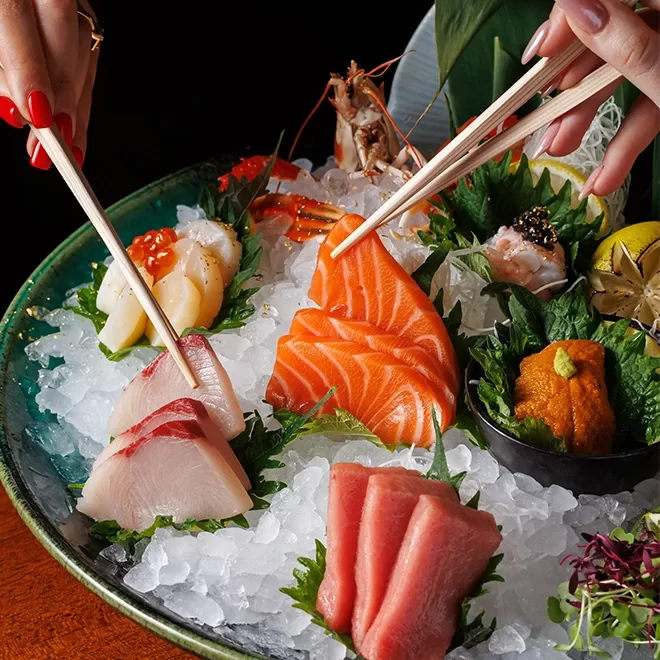 A colorful sashimi platter on ice with slices of salmon, tuna, yellowtail, scallop, uni, roe, and garnishes. Two hands with chopsticks are picking pieces from the arrangement.