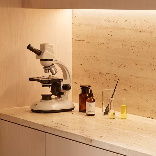 A white microscope sits on a beige marble countertop next to brown glass bottles, a glass beaker with a black dropper, and a tall glass containing yellow liquid, against a light wood and stone wall.