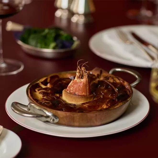 A copper dish filled with baked seafood pie, topped with a whole prawn standing upright in the center. The dish sits on a white plate with silverware and a glass of wine nearby on a red tablecloth.