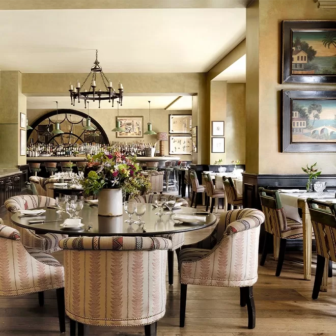 Elegant restaurant interior with patterned upholstered chairs, neatly set tables with glassware, a large flower arrangement centerpiece, paintings on the walls, and a well-stocked bar in the background. Warm and inviting atmosphere.