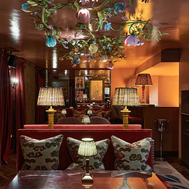 A warmly lit lounge with patterned pillows on red sofas, table lamps with striped shades, and a decorative ceiling fixture featuring colorful flowers and vines. The ambiance is cozy and eclectic.