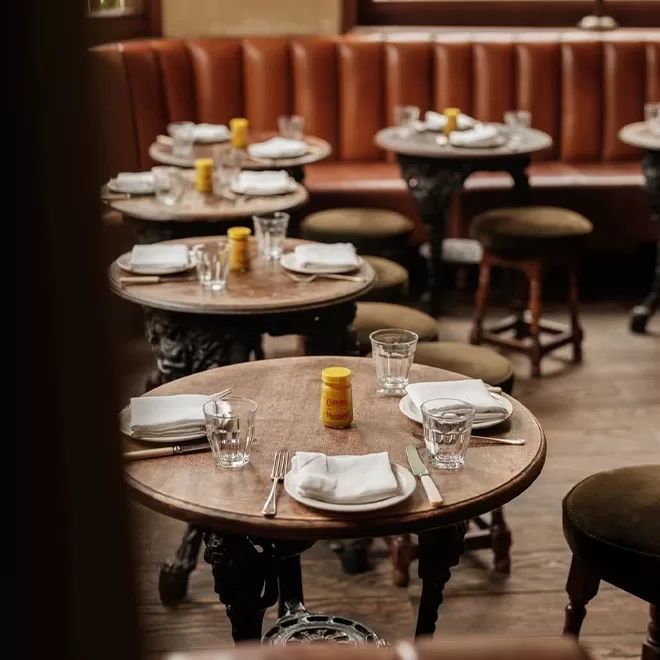 A cozy restaurant with round wooden tables set with glasses, white napkins, cutlery, and jars of mustard. Brown leather banquette seating lines the wall, and stools surround the tables. The atmosphere appears warm and inviting.