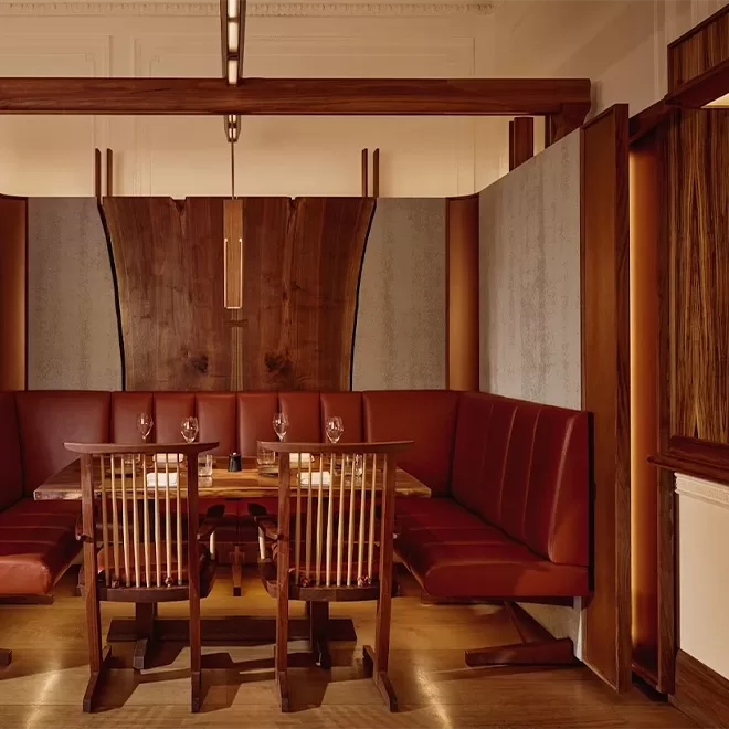 A cozy restaurant dining area with a wooden table, wine glasses set for four, wooden chairs, and a curved red upholstered bench, surrounded by wood and textured panel walls in warm lighting.