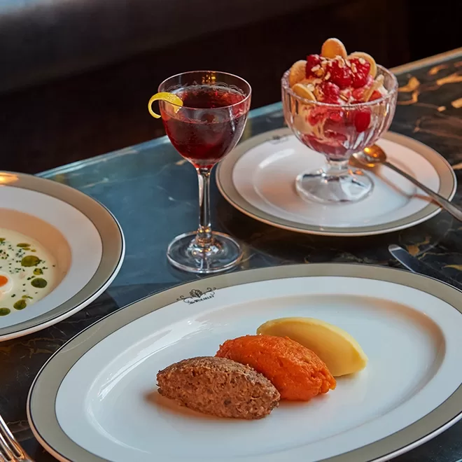 A table set with a plated meal: a meat quenelle, orange puree, and yellow puree on a white dish; a clear bowl of fruit dessert; a glass of red drink with a lemon twist; and a bowl of creamy soup with green oil.