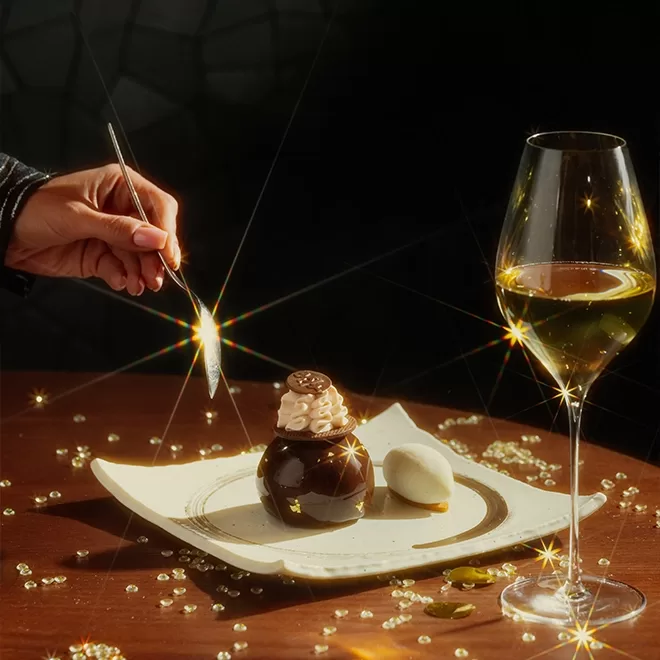 A hand holds a spoon above a fancy chocolate dessert on a plate, with a scoop of ice cream beside it. A glass of white wine and sparkling decorative gems are on the wooden table, illuminated by bright, star-like lights.