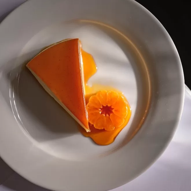 Slice of cheesecake with orange glaze on a white plate, decorated with orange sauce resembling a flower. Shadows create a striped pattern across the plate, enhancing the desserts presentation at Sessions Arts Club, Farringdon.