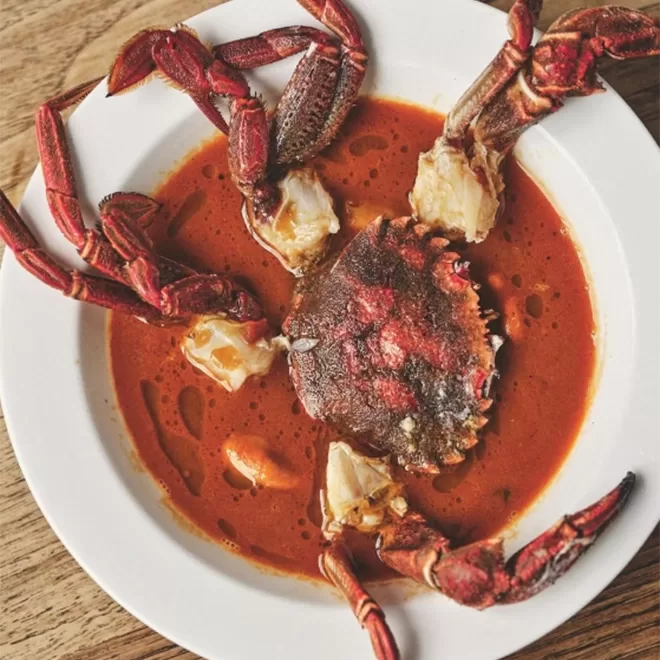 A vibrant stew displayed in a white bowl features a whole crab in a rich red broth. The crabs claws and legs are prominently arranged around its body, and the dish is set on a wooden table surface at BRAT Restaurant, Shoreditch.