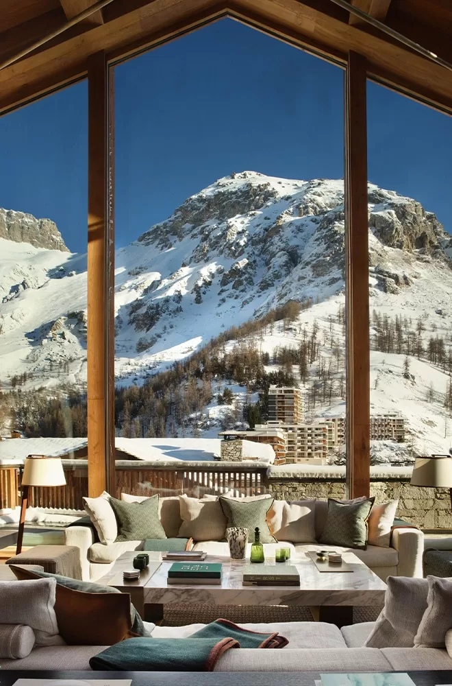 A cosy living room with large windows showcasing a breathtaking view of snowy mountains under a clear blue sky. The room features comfortable seating, a coffee table with books, and soft lighting, creating a warm atmosphere at Etoile du Nord, Val d’Isère.