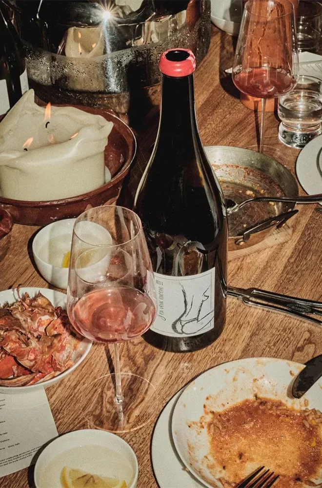 A bottle of wine with a red cap sits on a wooden table surrounded by glasses, a frying pan, candles, and plates with food remnants. Spears and shells are visible, suggesting a finished meal. Several glasses hold pink-hued liquid  at Mountain Beak Street, Soho, London.