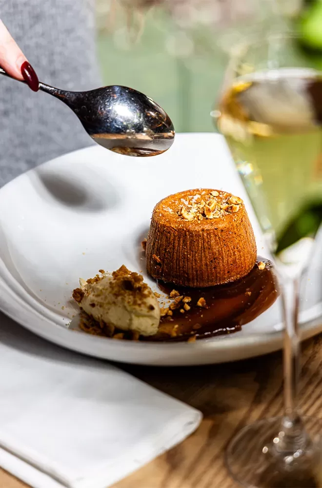 A person holds a spoon above a dessert, which appears to be a cake with caramel sauce and sprinkled nuts, served alongside a scoop of cream. A glass of white wine is nearby on a wooden table with a white napkin at Sucre, Soho, London.