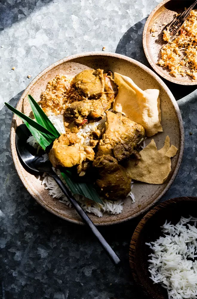 A plate of rice topped with pieces of chicken curry, garnished with green leaves and accompanied by poppadoms and coconut sambal. A spoon is placed on the plate, and a wooden bowl of rice is beside it at Kolamba, Soho, London.
