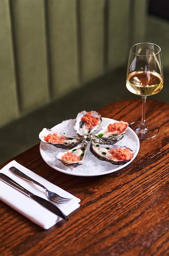 A plate of fresh oysters on ice, topped with a red garnish, on a wooden table beside a glass of white wine. A fork and knife are placed on a napkin next to the plate. The background has green upholstery at Donia, Soho, London.