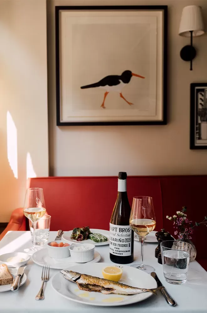 A table set for a meal with a grilled fish, lemon garnish, and side dishes. A bottle of wine and two glasses are present. The background features a framed bird illustration above a red cushioned bench. The scene is warmly lit at Quo Vadis, Soho, London.