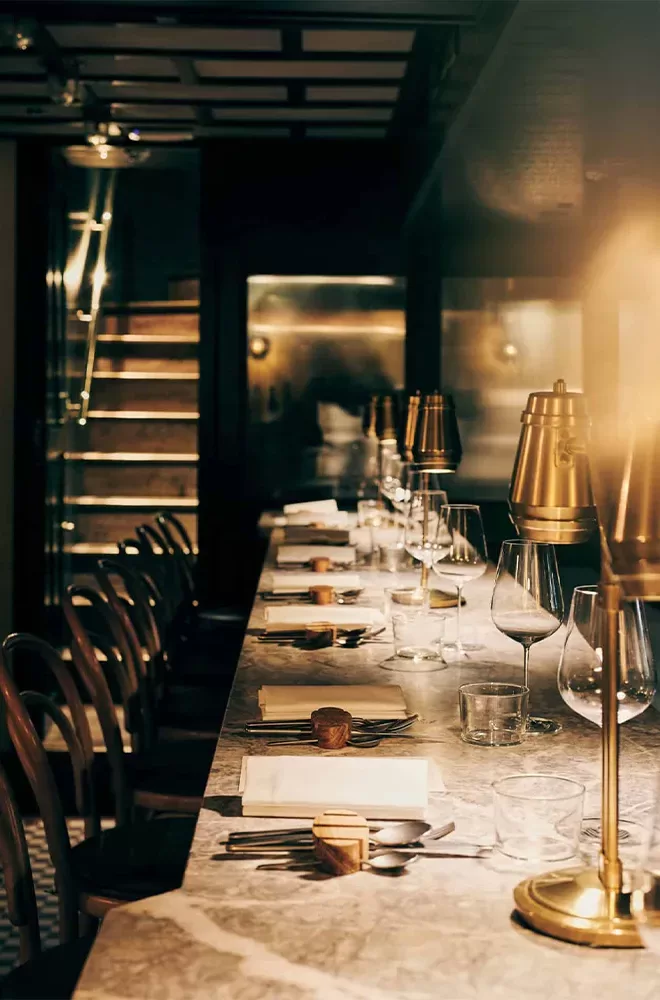 A dimly lit upscale restaurant bar with a marble countertop, set with empty wine glasses, napkins, and small lamps. Wooden bar stools line the counter, and a staircase is visible in the background. The ambiance is warm and inviting at Evelyn’s Table, Soho, London.