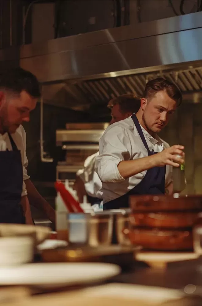 Chefs working in a restaurant kitchen. One chef is focused on seasoning a dish, while others prepare food in the background. The kitchen is busy, with various cooking utensils and ingredients visible on the counters at Mountain Beak Street, Soho, London.