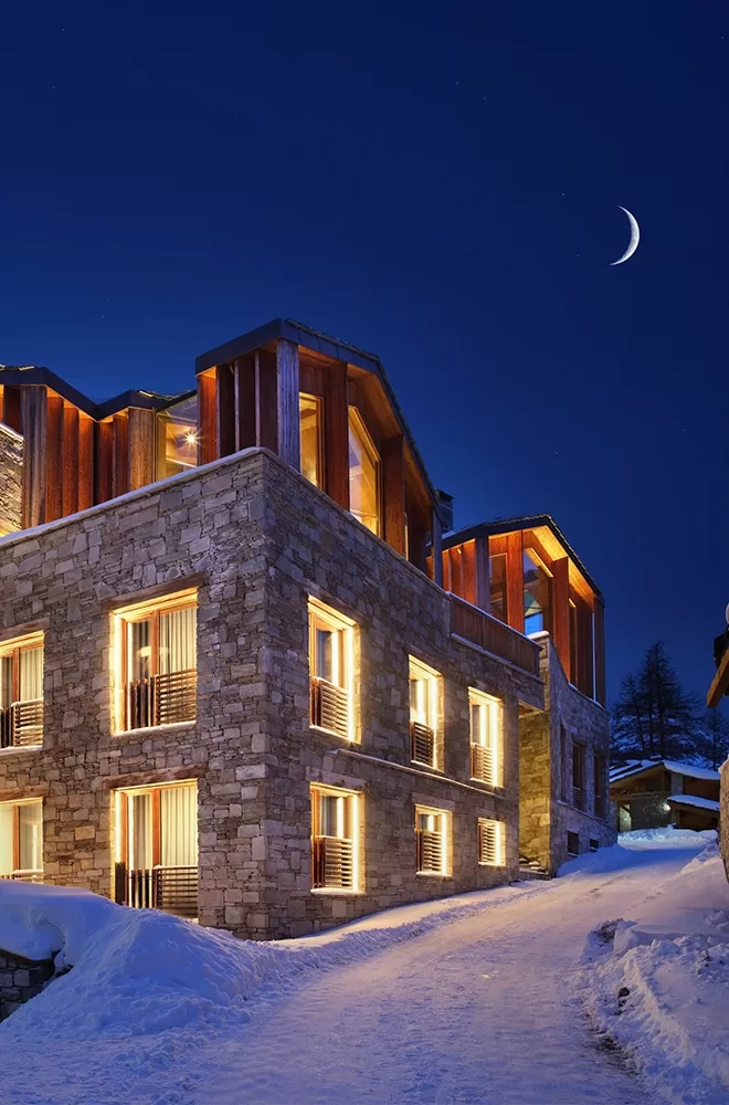A modern stone building glows warmly against a snowy landscape at night. The building features large windows and wooden accents. A crescent moon hangs in the clear, starry sky above Etoile du Nord, Val d’Isère.