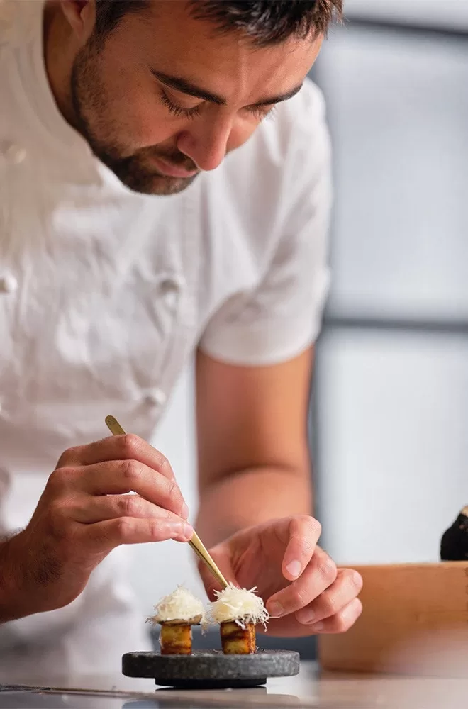 A chef in a white uniform delicately garnishes two small dishes with white toppings using tweezers. The dishes are placed on a dark platter. The background is softly blurred, focusing on the chefs precise movements at Aulis London, London.