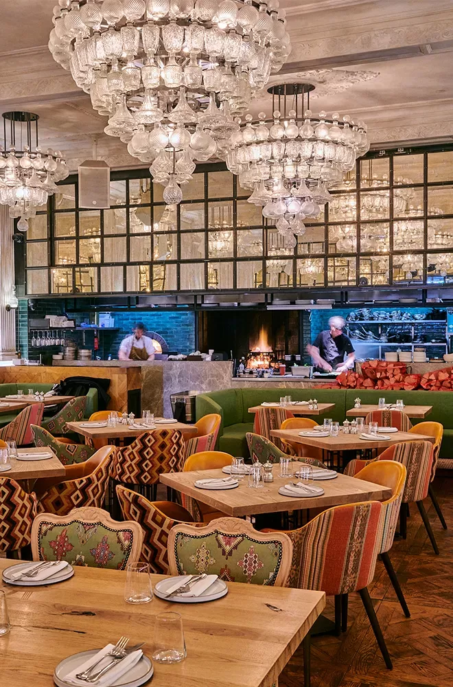 A stylish restaurant interior with ornate chandeliers, wooden tables set for dining, and colorful upholstered chairs. Two chefs are at work in an open kitchen with a wood-fired oven in the background at Sucre, Soho, London.