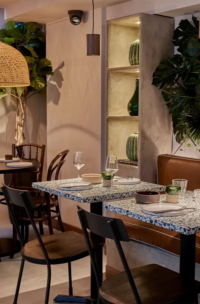 A cozy restaurant interior featuring wooden chairs and tables with terrazzo tops. Decorative plants and soft lighting from a woven lampshade create a warm atmosphere. Shelves display vases and greenery, adding to the inviting ambiance at Kolamba, Soho, London.