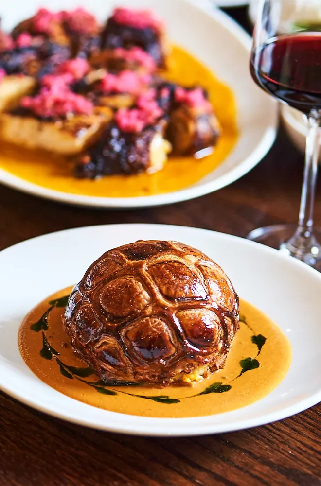 A gourmet dish featuring a round, browned pastry with a decorative pattern on top, served on a plate with a light brown sauce. In the background is another dish garnished with vibrant pink toppings. A glass of red wine is partially visible at Donia, Soho, London.