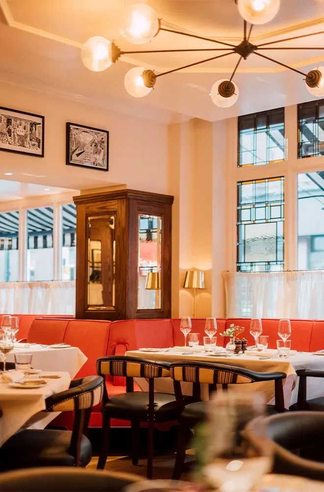 Cosy restaurant interior with red booth seating, elegantly set tables, and black chairs. Warm lighting from a chandelier illuminates the room. Framed art on the walls and decorative stained glass windows enhance the inviting ambience at Quo Vadis, Soho, London.