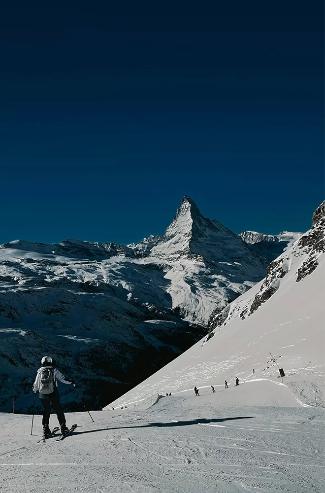 A skier descends a snowy slope under a clear blue sky, with the iconic Matterhorn mountain in the background. The scene captures the serene and majestic winter landscape at Mont Cervin Palace, Zermatt.
