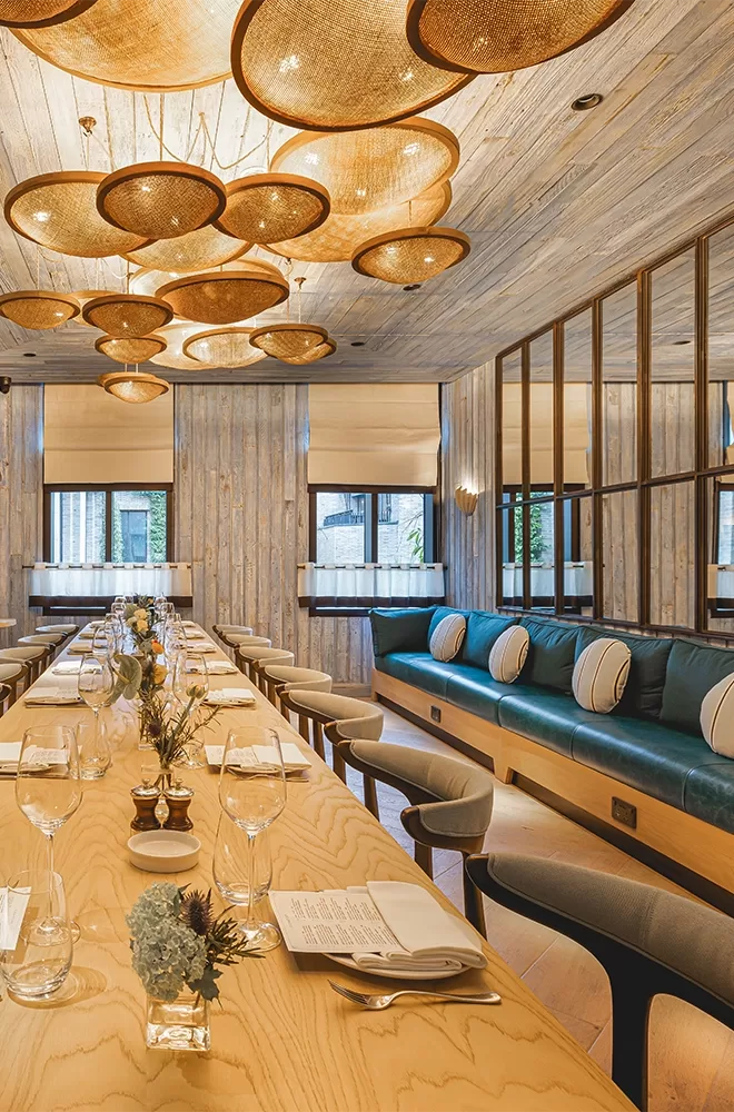 A stylish dining area with a long wooden table set for a meal. Modern chairs and cushioned bench seating line the sides. Above, unique circular pendant lights hang, and natural light flows in through windows with blinds at Azzurra, London.