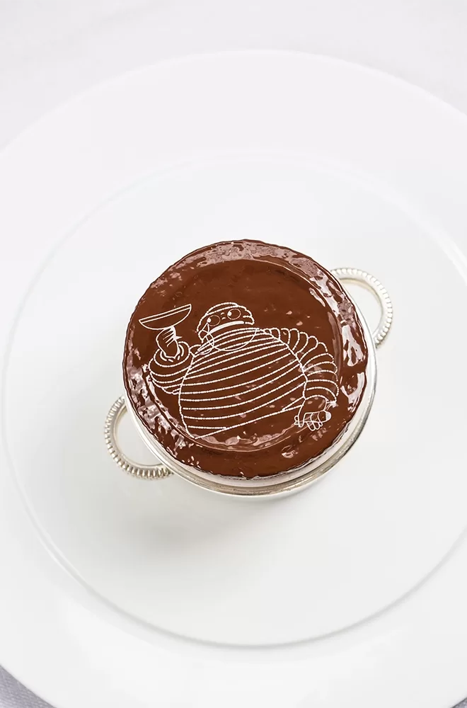 A chocolate dessert on a white plate, featuring an illustration of a cartoonish, round character holding a drink etched into the chocolate surface. The dessert is placed in a silver dish with handles at Claude Bosi at Bibendum.
