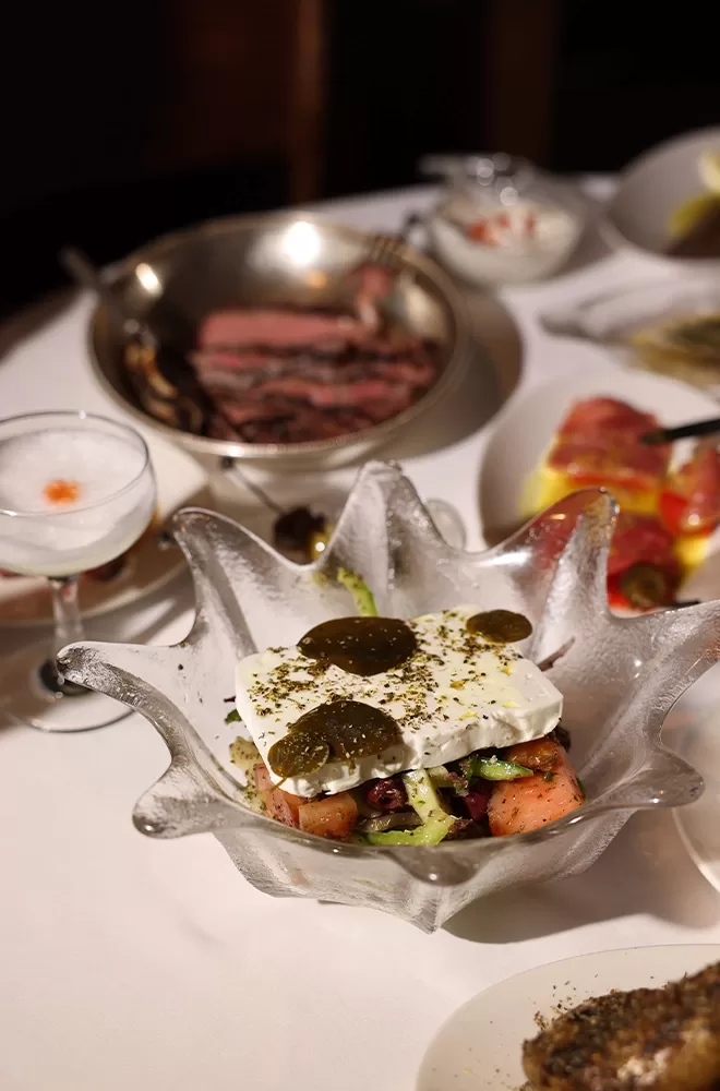 A table set with various dishes, including a salad in a decorative glass bowl topped with feta cheese and herbs, a dish of sliced meat, a cocktail, and other appetizers on white plates. The setting suggests a fine dining experience at Bottarga, London.