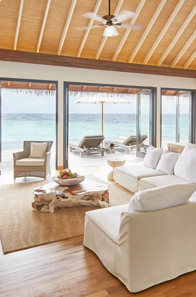 A bright, airy living room with white sofas, a rustic wooden coffee table, and a bowl of fruit. Large glass doors open to a deck with lounge chairs, offering a view of the ocean. The room has a high, wooden ceiling with a ceiling fan at COMO Maalifushi, Thaa Atoll, Maldives.