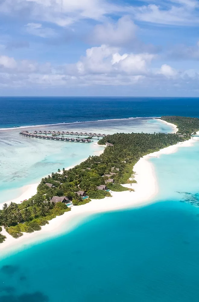 Aerial view of a tropical island surrounded by turquoise waters. There are overwater bungalows extending into the ocean, lush green vegetation, and white sandy beaches under a partly cloudy sky at Niyama, Dhaalu Atoll, Maldives.