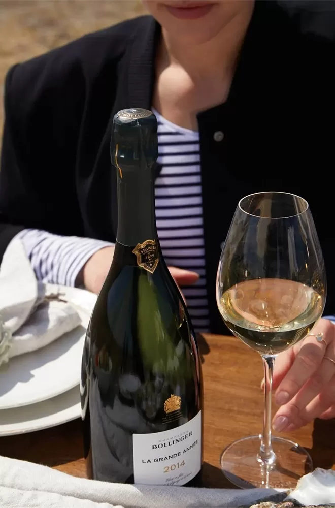 A person sitting at a table holds a glass of white wine next to a bottle of Bollinger La Grande Année 2014 Champagne. A plate and napkin are in the background. The person wears a striped shirt and dark jacket.