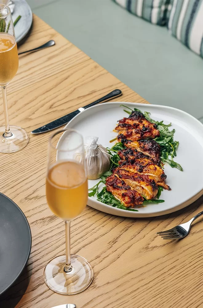 A plate of grilled chicken served on a bed of greens with a wrapped lemon, placed on a wooden table. Two glasses of orange-colored beverages are nearby, along with cutlery. The table has a light wood finish and features striped cushions in the background at Azzurra, London.